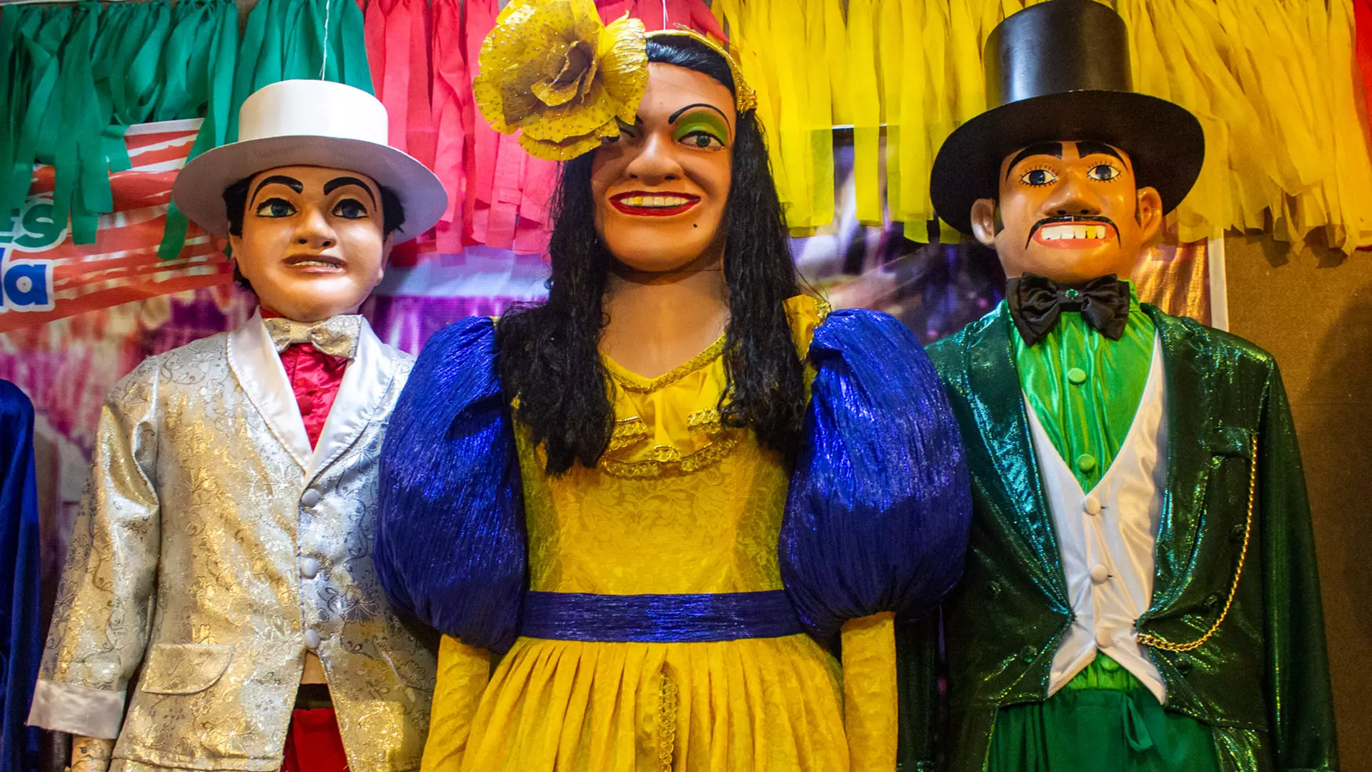 Três bonecos gigantes coloridos com roupas festivas, posando em frente a bandeirinhas de Carnaval.